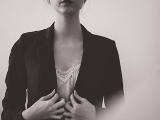 A monochrome portrait of a person wearing a dark blazer over a light, lace-trimmed top. The hands are gently holding the blazer, and the lighting creates a soft, subdued atmosphere.