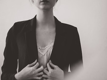 A monochrome portrait of a person wearing a dark blazer over a light, lace-trimmed top. The hands are gently holding the blazer, and the lighting creates a soft, subdued atmosphere.