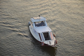 A small white motorboat is navigating through calm waters. The boat has a covered cabin with windows, life buoys attached to the sides, and a small antenna on top. The water reflects a golden hue under the sunlight.