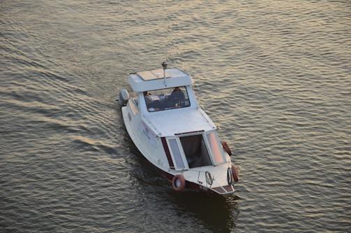 A small white motorboat is navigating through calm waters. The boat has a covered cabin with windows, life buoys attached to the sides, and a small antenna on top. The water reflects a golden hue under the sunlight.