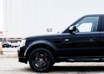 A close-up view of a black SUV parked in front of a large white trailer with the number 53' visible. The scene takes place in an industrial area or parking lot with other vehicles and a building partially visible in the background.