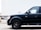 A close-up view of a black SUV parked in front of a large white trailer with the number 53' visible. The scene takes place in an industrial area or parking lot with other vehicles and a building partially visible in the background.