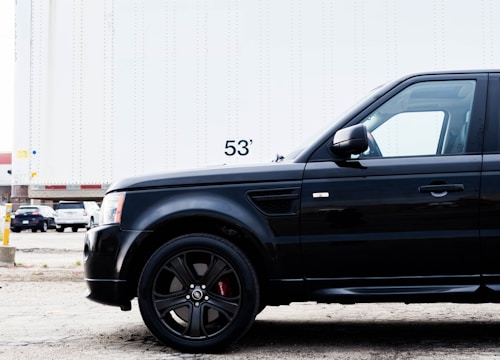 A close-up view of a black SUV parked in front of a large white trailer with the number 53' visible. The scene takes place in an industrial area or parking lot with other vehicles and a building partially visible in the background.
