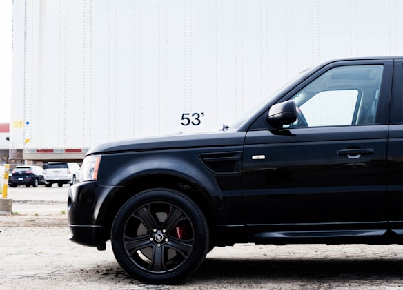 A close-up view of a black SUV parked in front of a large white trailer with the number 53' visible. The scene takes place in an industrial area or parking lot with other vehicles and a building partially visible in the background.