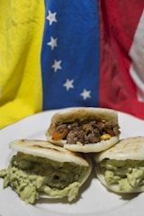 Three arepas filled with different ingredients sit on a white plate. One is filled with shredded beef and vegetables like corn and tomatoes, while the other two are filled with a creamy, green avocado mixture. The background features the flag of Venezuela, with sections of yellow, blue, and red that have white stars on the blue stripe.