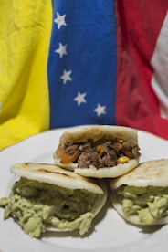 Three arepas filled with different ingredients sit on a white plate. One is filled with shredded beef and vegetables like corn and tomatoes, while the other two are filled with a creamy, green avocado mixture. The background features the flag of Venezuela, with sections of yellow, blue, and red that have white stars on the blue stripe.