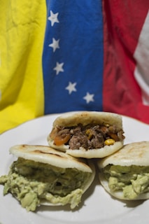 Three arepas filled with different ingredients sit on a white plate. One is filled with shredded beef and vegetables like corn and tomatoes, while the other two are filled with a creamy, green avocado mixture. The background features the flag of Venezuela, with sections of yellow, blue, and red that have white stars on the blue stripe.
