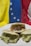 Three arepas filled with different ingredients sit on a white plate. One is filled with shredded beef and vegetables like corn and tomatoes, while the other two are filled with a creamy, green avocado mixture. The background features the flag of Venezuela, with sections of yellow, blue, and red that have white stars on the blue stripe.