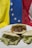 Three arepas filled with different ingredients sit on a white plate. One is filled with shredded beef and vegetables like corn and tomatoes, while the other two are filled with a creamy, green avocado mixture. The background features the flag of Venezuela, with sections of yellow, blue, and red that have white stars on the blue stripe.