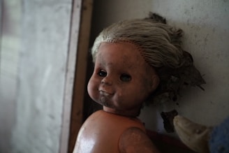 A vintage doll with a worn and aged appearance is leaning against a wall. It has pale, disheveled hair and a cracked, dirty face. The eyes appear hollow, adding a haunting quality. The background is dimly lit, contributing to the eerie atmosphere.