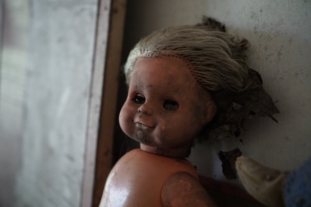 A vintage doll with a worn and aged appearance is leaning against a wall. It has pale, disheveled hair and a cracked, dirty face. The eyes appear hollow, adding a haunting quality. The background is dimly lit, contributing to the eerie atmosphere.