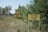 A warning sign with a radiation symbol next to a sign with Cyrillic script is placed in a grassy, wooded area. The background features sparse trees with green foliage and a railway track running through the scene.