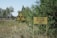 A warning sign with a radiation symbol next to a sign with Cyrillic script is placed in a grassy, wooded area. The background features sparse trees with green foliage and a railway track running through the scene.