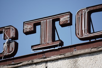 Rusted metal letters on top of a building feature a vintage sign. The letters are accented with white tubing, creating a contrast against the blue sky background. The structure appears weathered, with peeling and cracked surfaces.