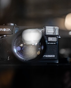 A vintage camera with the brand name partially visible, featuring a large lens and a flash component. The image is focused on the camera's lens, and the background appears blurred, creating an artistic effect.