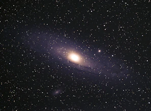 A vast expanse of space filled with numerous stars scattered across the dark sky. At the center, a glowing galaxy with a bright core and spiraling arms is prominent, showcasing the grand scale of the universe.