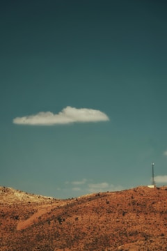 A single white cloud floats in a vast, clear blue sky above a barren, reddish-brown landscape that appears to be dry and rocky. On the right side of the horizon stands a tall communication tower, adding a touch of technology to the otherwise natural scene.