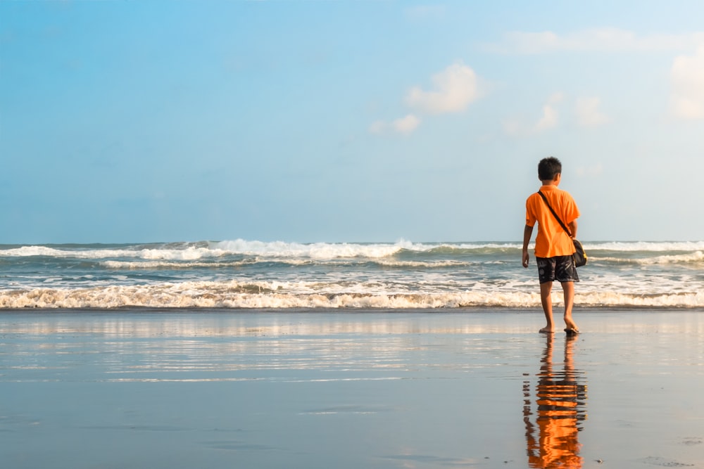 man in orange shirt and black shorts walking on seashore during daytime