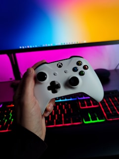 A hand is holding a white gaming controller in front of a computer setup. The background features a monitor displaying a colorful gradient, along with a keyboard lit with vibrant RGB lighting.