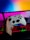 A hand is holding a white gaming controller in front of a computer setup. The background features a monitor displaying a colorful gradient, along with a keyboard lit with vibrant RGB lighting.