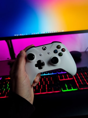 A hand is holding a white gaming controller in front of a computer setup. The background features a monitor displaying a colorful gradient, along with a keyboard lit with vibrant RGB lighting.