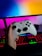 A hand is holding a white gaming controller in front of a computer setup. The background features a monitor displaying a colorful gradient, along with a keyboard lit with vibrant RGB lighting.
