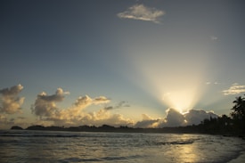 Sunrise over a tranquil coastal landscape with rays of light streaming through clouds. Gently rippling ocean water in the foreground and silhouetted distant landmasses and palm trees add to the serene scene.