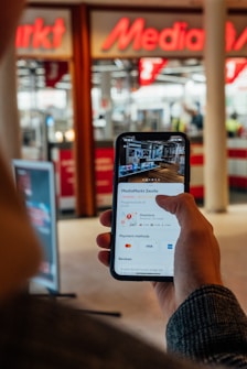 A person holds a smartphone displaying information about a store. The store's name, location, directions, payment methods, and reviews are visible on the screen. The background shows a blurred view of a store entrance with bright signage.