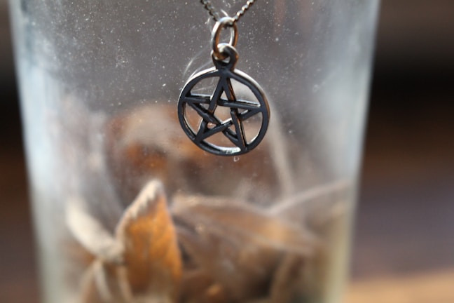 A necklace with a pentagram pendant is positioned inside a transparent glass, while dried leaves are scattered at the bottom. The image has a soft focus and subdued lighting.