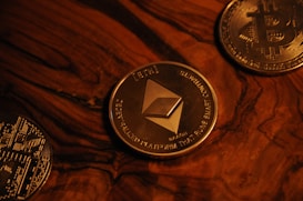 Three cryptocurrency coins on a wooden surface include Ethereum in the center, surrounded by Bitcoin coins. The coins are metallic with detailed engravings and symbols characteristic of blockchain technology.