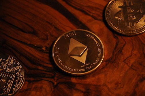 Three cryptocurrency coins on a wooden surface include Ethereum in the center, surrounded by Bitcoin coins. The coins are metallic with detailed engravings and symbols characteristic of blockchain technology.