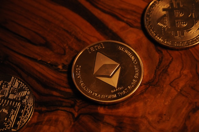 Three cryptocurrency coins on a wooden surface include Ethereum in the center, surrounded by Bitcoin coins. The coins are metallic with detailed engravings and symbols characteristic of blockchain technology.