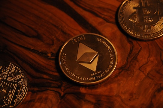 Three cryptocurrency coins on a wooden surface include Ethereum in the center, surrounded by Bitcoin coins. The coins are metallic with detailed engravings and symbols characteristic of blockchain technology.