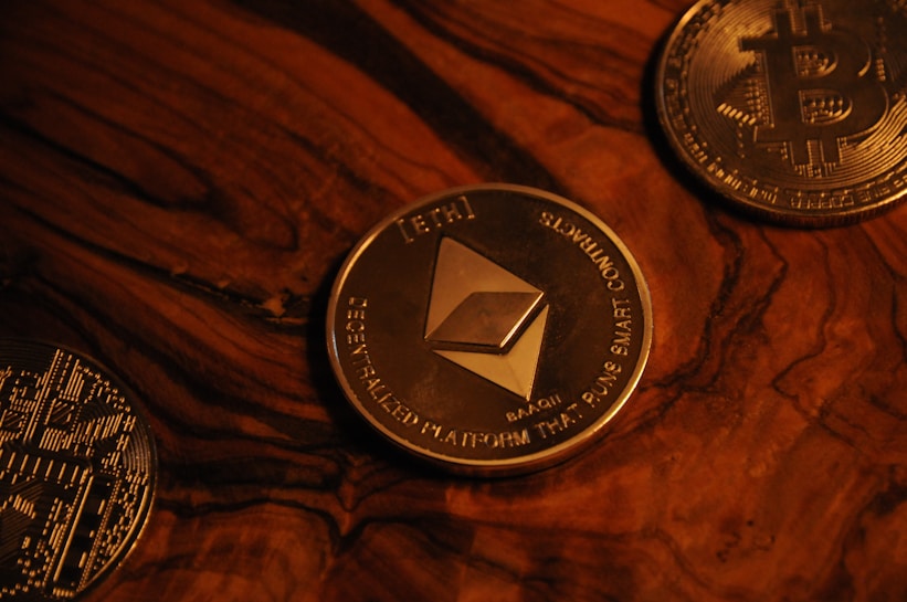 Three cryptocurrency coins on a wooden surface include Ethereum in the center, surrounded by Bitcoin coins. The coins are metallic with detailed engravings and symbols characteristic of blockchain technology.