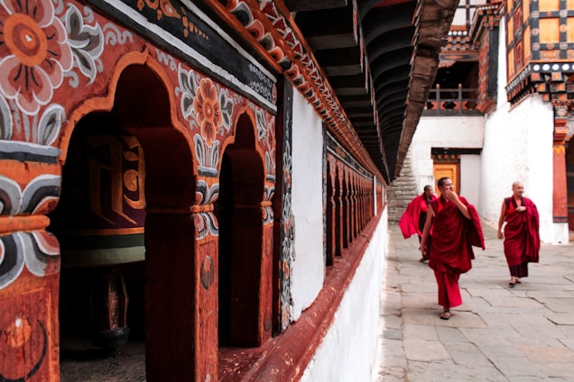 Monks in maroon robes walk past golden prayer bells in a serene Bhutanese monastery setting
