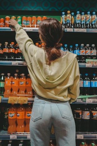 A person wearing a light green hoodie and jeans is reaching for a can on the top shelf in a grocery store aisle. The shelves are stocked with various soft drink bottles, including Fanta and Pepsi. The lighting is warm and enhances the vibrant colors of the products.