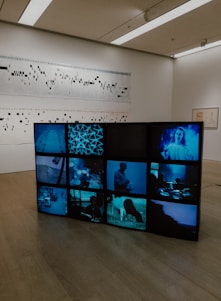 A contemporary art exhibit in a gallery featuring a video installation composed of multiple screens displaying different scenes, predominantly in shades of blue. Behind the video installation, there is a wall with abstract artwork resembling a graph or musical score.