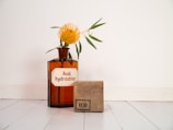 A brown glass bottle labeled 'Acid. hydrochlor.' is placed on a white surface, containing a yellow flower with green leaves. Next to it, a rectangular stone or block with black markings is positioned.