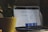 A MacBook Pro laptop displaying a webpage with text and small profile images. It sits on a dark surface with part of a large green potted plant visible to the left side. The setting appears to be indoors with dim lighting, creating a cozy atmosphere.