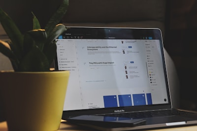 A MacBook Pro laptop displaying a webpage with text and small profile images. It sits on a dark surface with part of a large green potted plant visible to the left side. The setting appears to be indoors with dim lighting, creating a cozy atmosphere.