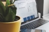 A laptop displaying a webpage with text and profiles alongside a green potted plant, creating a home-office vibe. The screen shows discussions on technical topics. A smartphone is partially visible near the laptop.