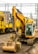 A large yellow excavator is positioned on a gravelly surface near a railway. Behind it, there are multiple yellow railway maintenance vehicles. Overhead power lines and a structure with a spire can be seen in the background, suggesting an urban environment.