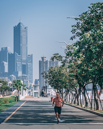 A shirtless man is jogging along a tree-lined pathway in an urban area, with skyscrapers and tall buildings in the background. The setting appears to be a clear day, with abundant greenery and modern streetlights.