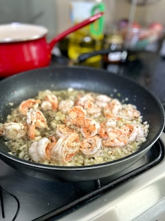 Shrimp are being cooked in a skillet with garlic and herbs, creating a savory mixture. The pan is positioned on a stove top, and the background features a red pot and some kitchen items, slightly blurred.
