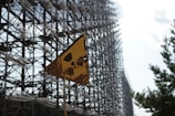 A rusted yellow triangular sign with a radiation symbol stands in front of a massive intricate metal structure. Trees are visible on the right side, and the sky is partially cloudy.