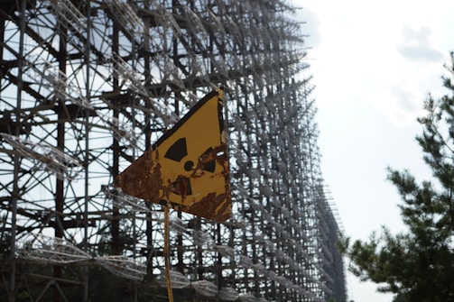 A rusted yellow triangular sign with a radiation symbol stands in front of a massive intricate metal structure. Trees are visible on the right side, and the sky is partially cloudy.