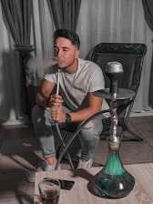 A person is sitting on a chair in a relaxed posture, exhaling smoke from a hookah pipe. The room has dark curtains and tiled flooring, with the hookah placed on a low wooden table in front of the individual. There is a drink in a glass on the table beside a smartphone.