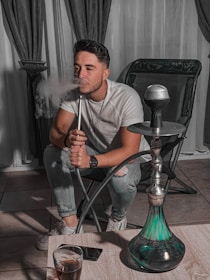 A person is sitting on a chair in a relaxed posture, exhaling smoke from a hookah pipe. The room has dark curtains and tiled flooring, with the hookah placed on a low wooden table in front of the individual. There is a drink in a glass on the table beside a smartphone.
