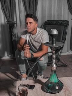 A person is sitting on a chair in a relaxed posture, exhaling smoke from a hookah pipe. The room has dark curtains and tiled flooring, with the hookah placed on a low wooden table in front of the individual. There is a drink in a glass on the table beside a smartphone.