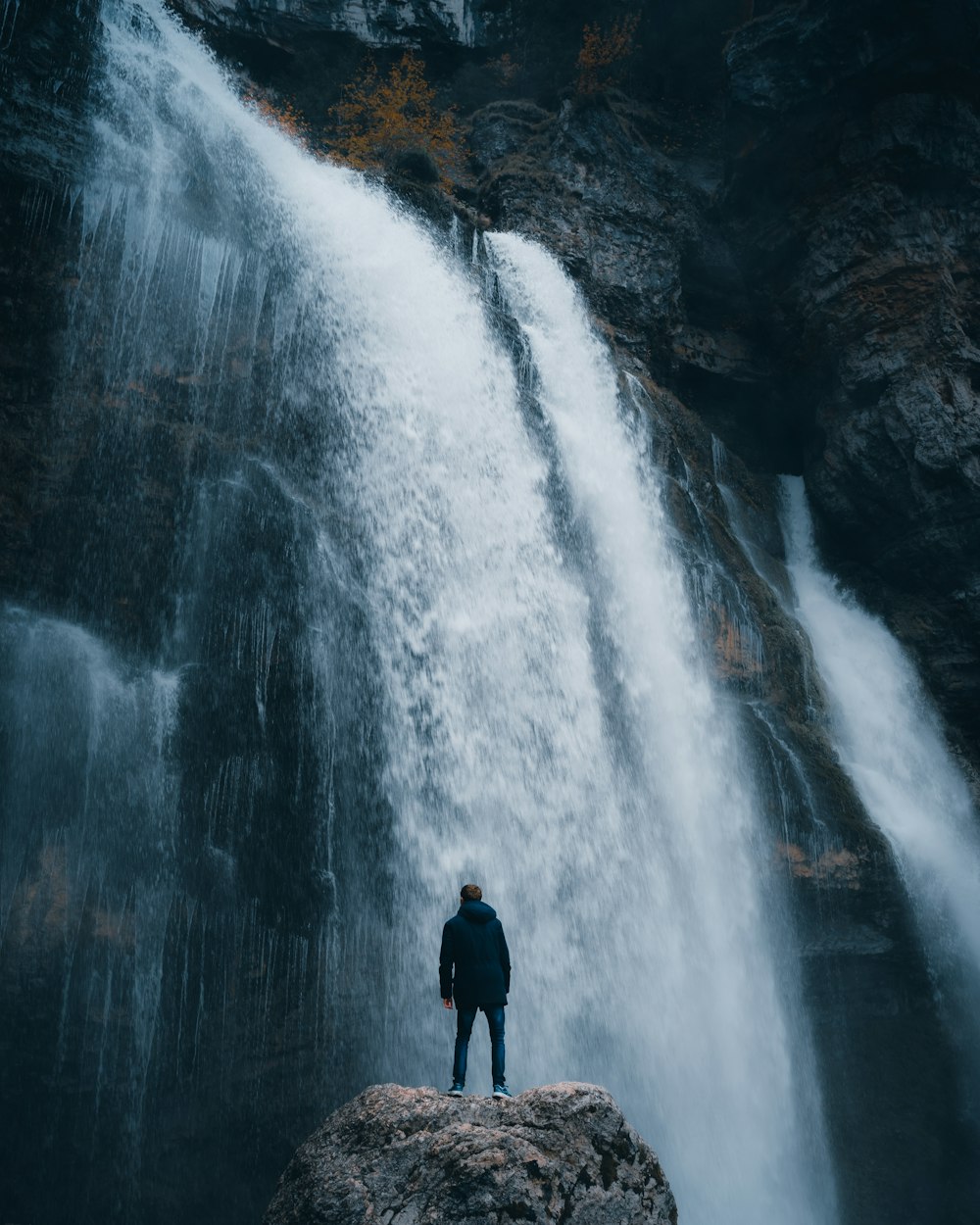 昼間、滝の近くの岩の上に立つ黒いジャケットの男